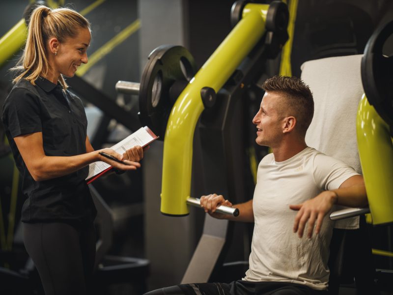 Shot of a muscular young woman in sportswear working out with personal trainer at the gym machine. She is pumping up her chest muscule with heavy weight.